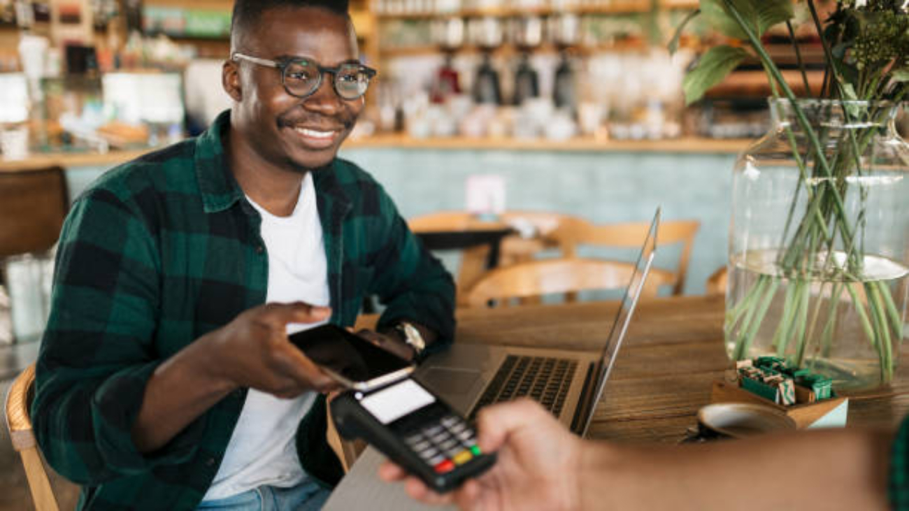 Cheerful and handsome young African American male student sitting in a cafe, working on his laptop and using his smart phone to pay for his drink, smiling